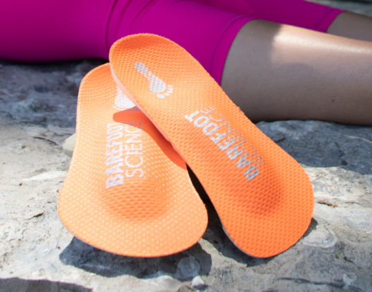 Orange 3/4 length textured insoles with Barefoot Science and Evergreen logos printed in white, resting on stone surface.