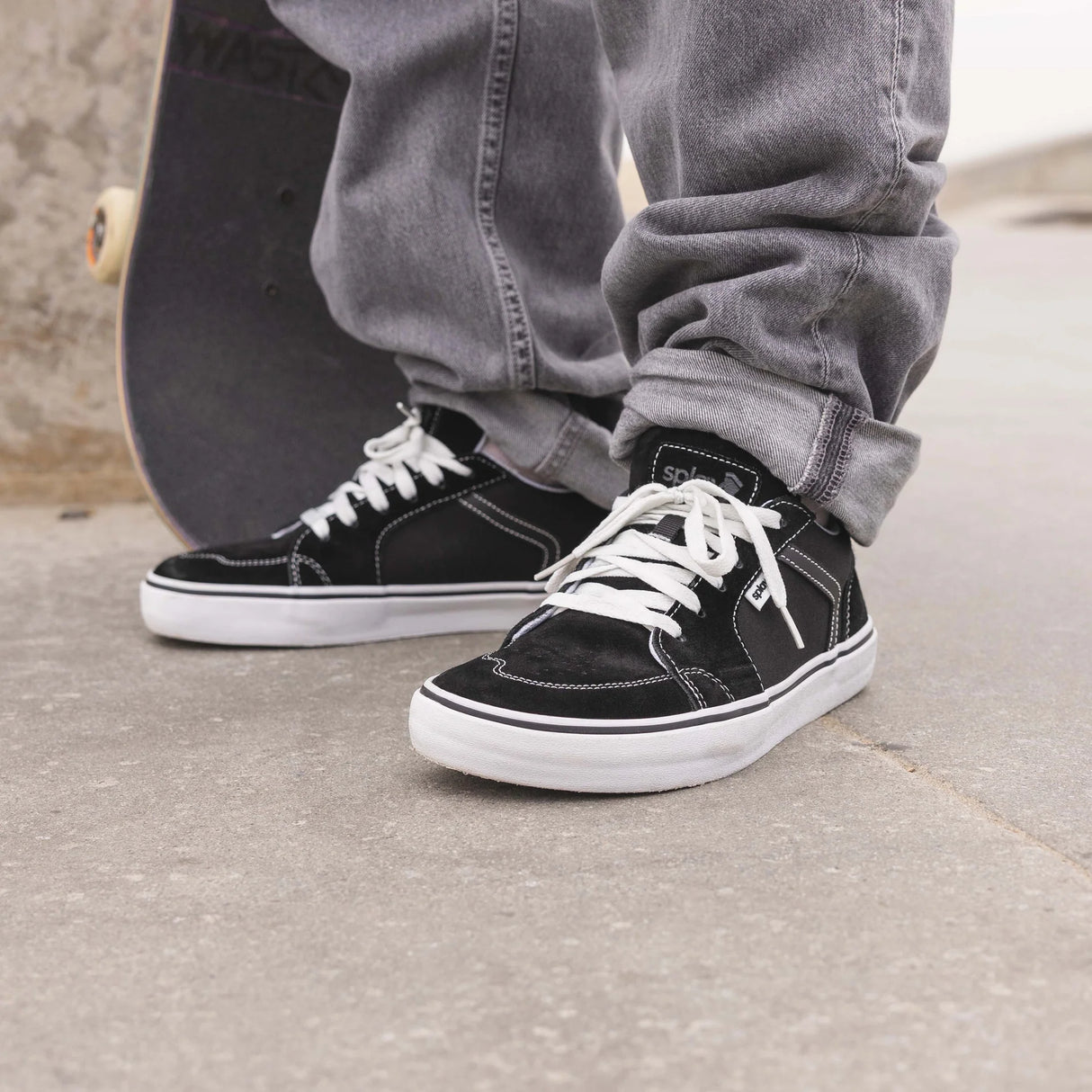Person wearing black sneakers with white laces on a concrete surface, with a skateboard in the background.