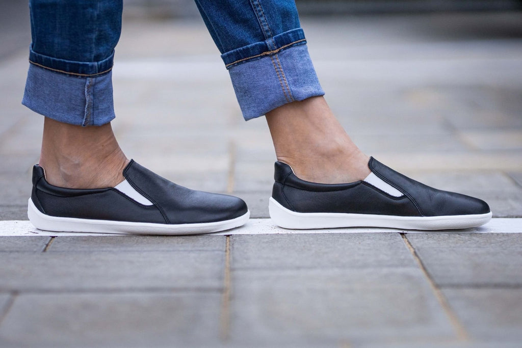 Black slip-on sneakers with white soles, smooth leather upper, and elastic side panels worn with cuffed jeans.
