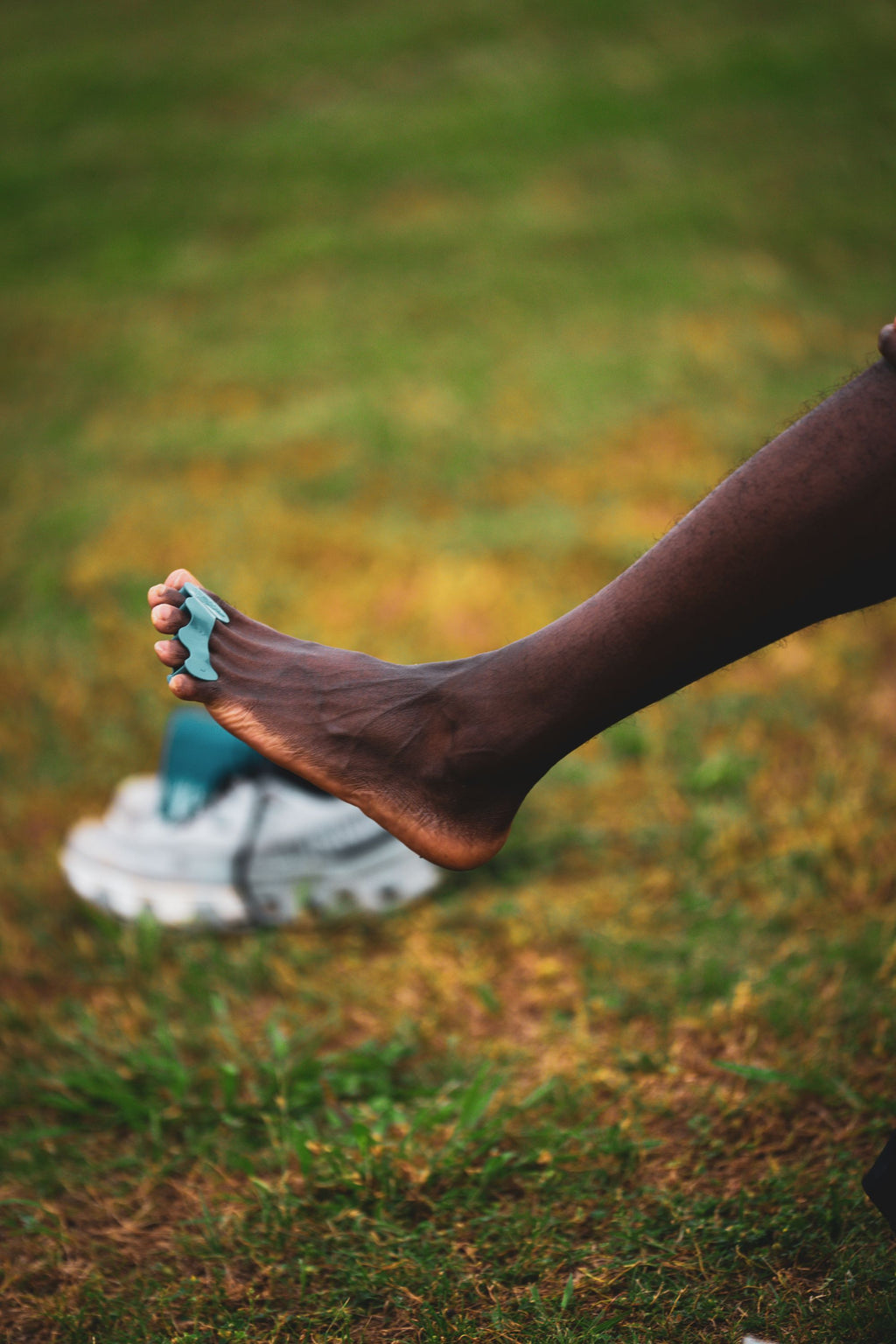 Dark-skinned foot wearing mauve toe spacers stretching outdoors over grass with gray shoes in background.