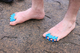 Feet wearing aqua blue Correct Toes® toe spacers on a rocky surface, showing separated toes.