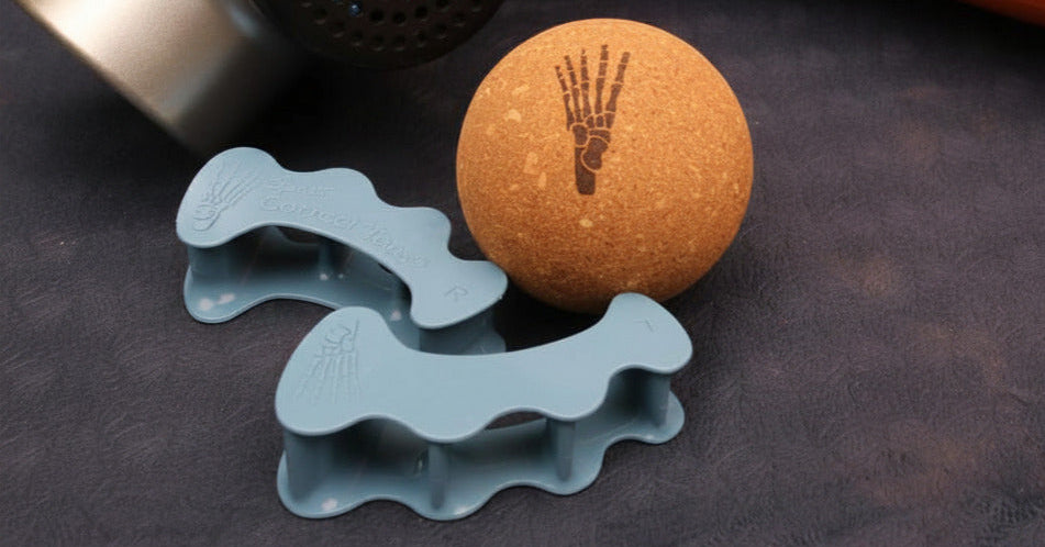 Massage gun, orange foam roller, cork massage ball with a handprint logo, and two blue silicone massage tools arranged on a dark textured surface.