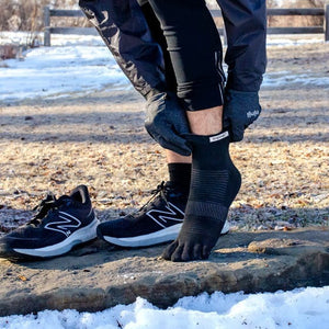 Black five-toe mini crew socks worn outdoors with black athletic shoes nearby on a snowy trail.