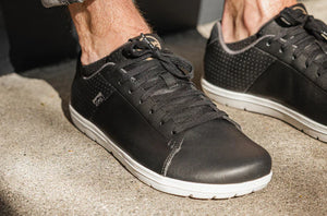 Black low-top casual sneaker with white sole and textured side panel, worn on foot outdoors.