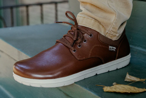 Brown leather casual shoe with white sole and round toe, worn with beige pants.