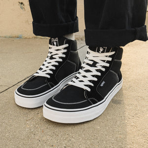 Black high-top sneakers with white laces worn by a person on a concrete surface.