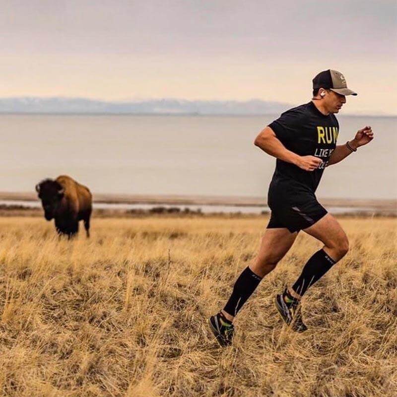 Man wearing black Zensah compression leg sleeves running through dry grassland with bison in the background.
