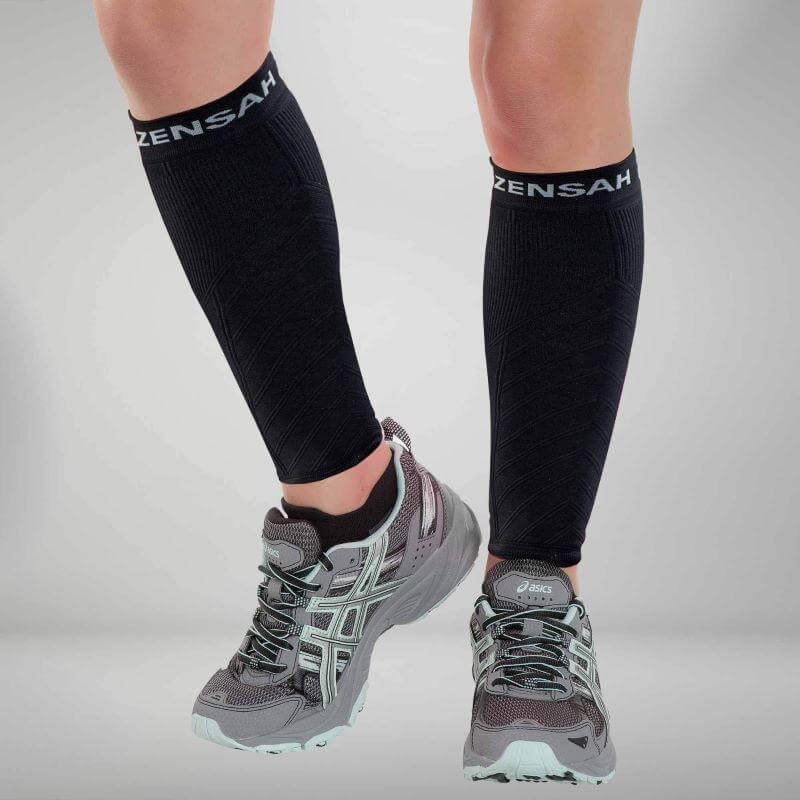 Black knee-high compression leg sleeves with white Zensah logo worn with gray running shoes.